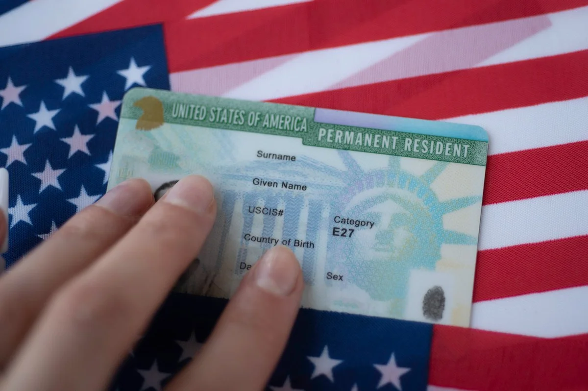 Foto colotrida de green card de residente permanente dos Estados Unidos, sobre a bandeira norte-americana - Metrópoles